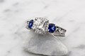 
Vintage Three Stone Scrollwork Engagement Ring in platinum with a 6mm cushion cut diamond center stone and cushion cut lab created blue sapphire side stones.