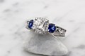 
Vintage Three Stone Scrollwork Engagement Ring in platinum with a 6mm cushion cut diamond center stone and cushion cut lab created blue sapphire side stones.