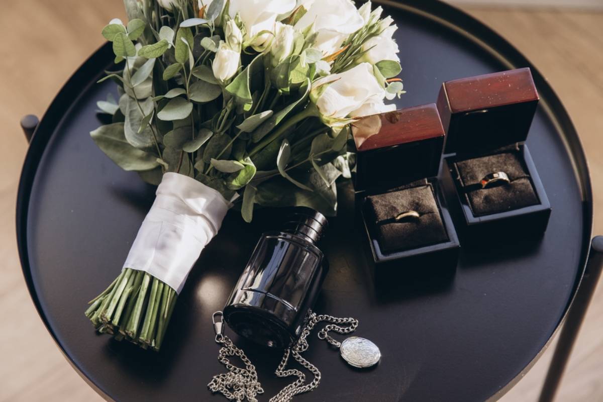 Wedding ring photos showing gold bands in boxes beside a bridal bouquet, perfume bottle, and locket on a black table.